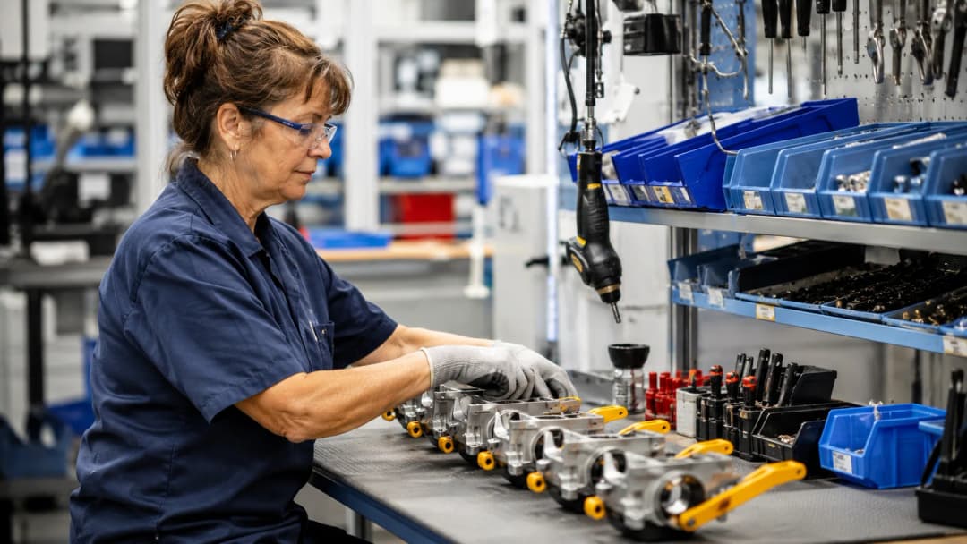 A woman assembles mechanical parts on a factory workbench with tools and parts bins nearby
