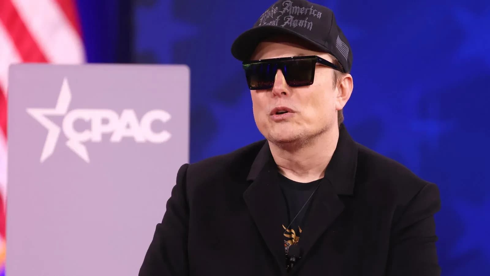 Elon Musk in a black cap and sunglasses speaking at the Conservative Political Action Conference