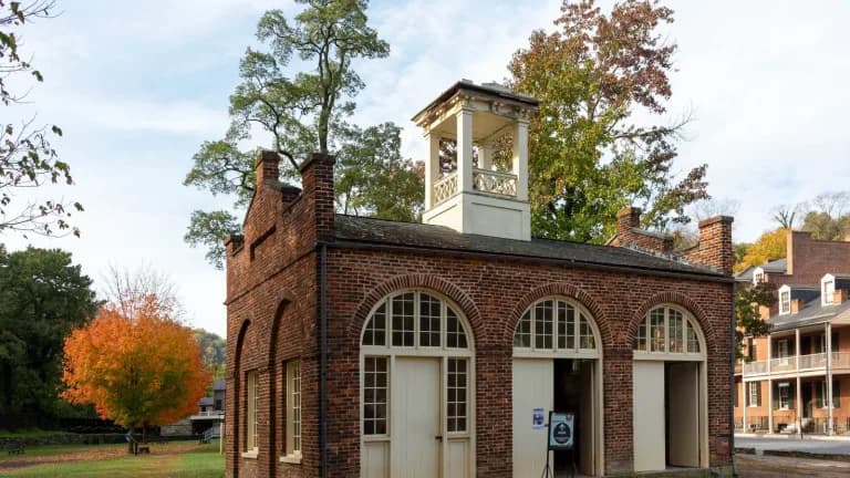 A small brick engine house with a white cupola and arched doors, framed by autumn trees