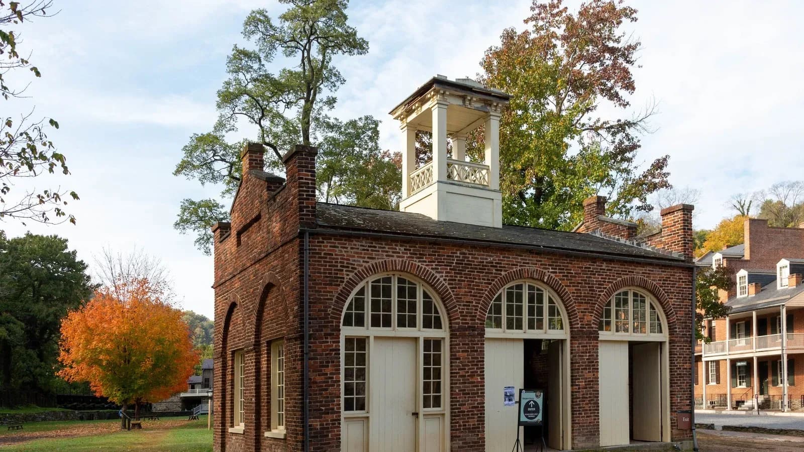 A small brick engine house with a white cupola and arched doors, framed by autumn trees
