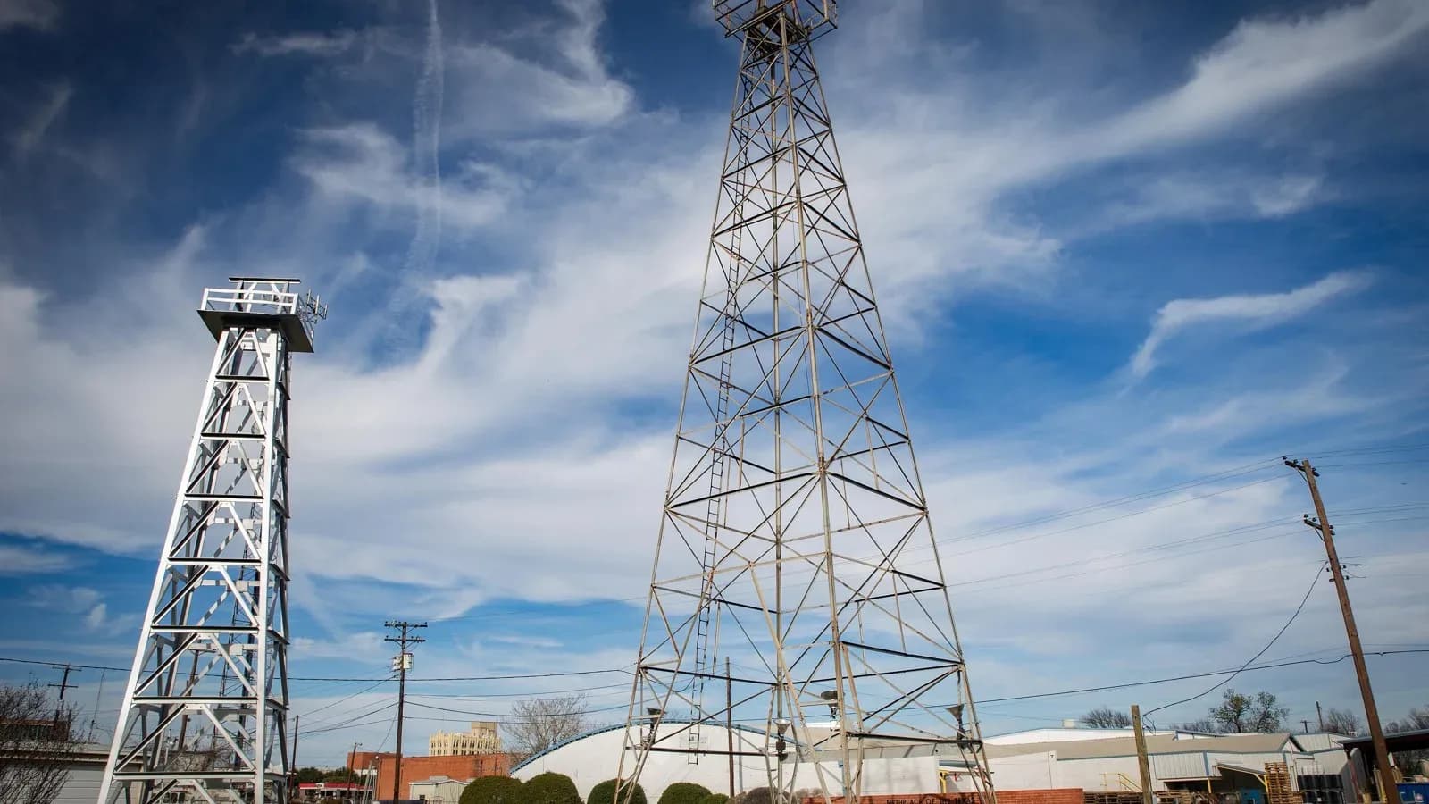 Two steel oil derricks, one white and one bare metal, rising against a blue sky with wispy clouds in Corsicana