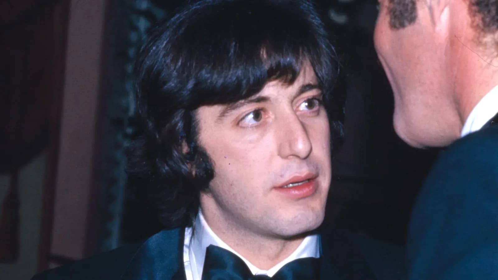 A young Al Pacino in a tuxedo and bow tie speaking with James Caan