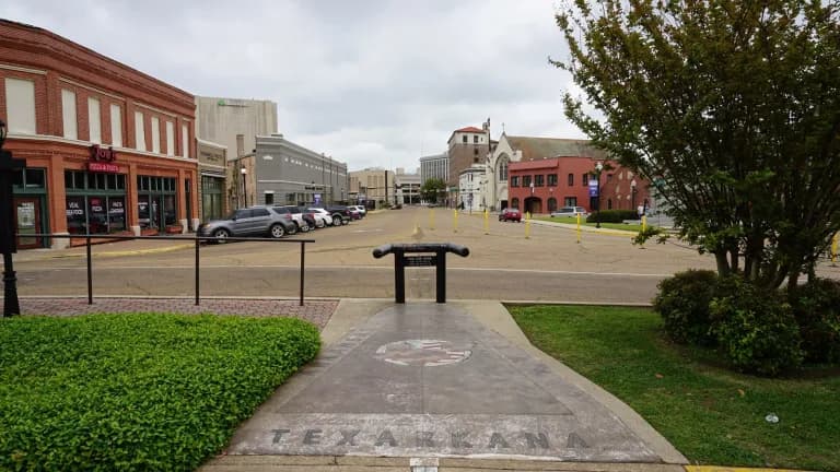 A sidewalk marker reading Texarkana on State Line Avenue, with brick downtown buildings in the background