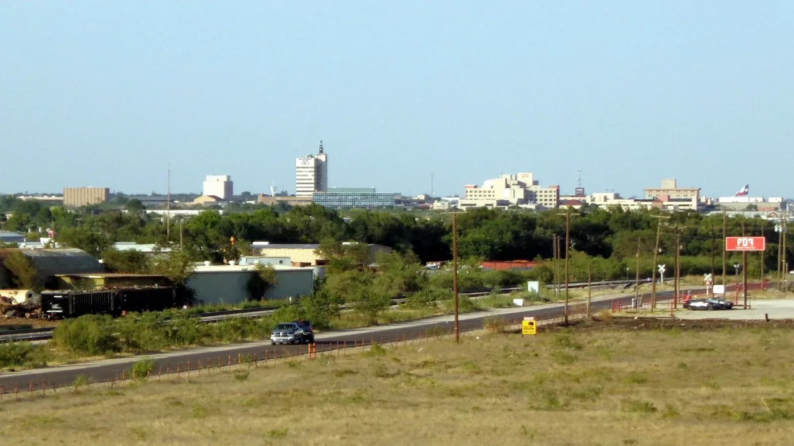 The downtown Odessa skyline seen across open grassland and a road under a clear blue sky