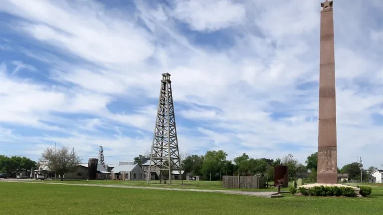 A wooden oil derrick and a tall granite monument on the lawn of the Spindletop-Gladys City Boomtown Museum under a blue sky