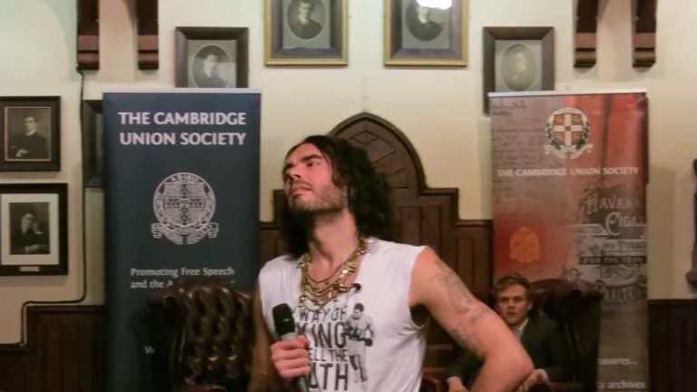 Russell Brand holding a microphone while speaking at the Cambridge Union Society