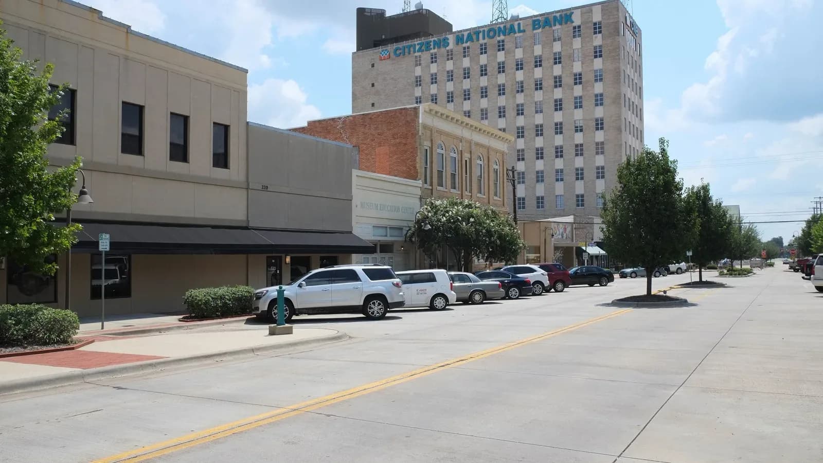 A wide street in downtown Longview lined with parked cars, low brick buildings and a tall office tower