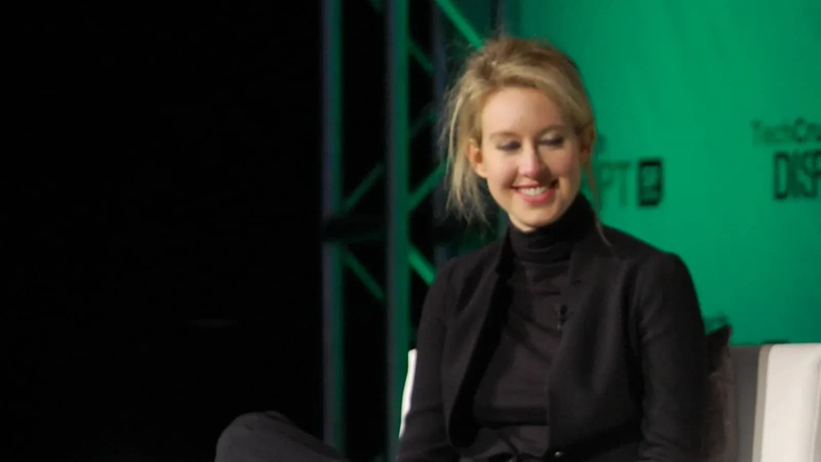 Elizabeth Holmes, smiling, seated onstage in a black jacket and turtleneck against a green backdrop