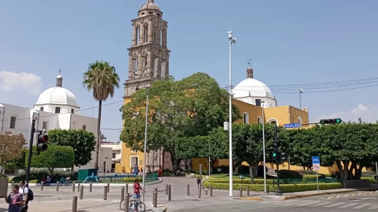 A colonial church bell tower and white domes rise above a tree-lined plaza in Irapuato, Mexico