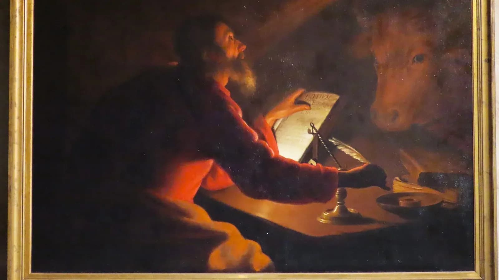 Candlelit painting of St. Luke writing at a desk, looking upward, with an ox, his traditional symbol, behind him