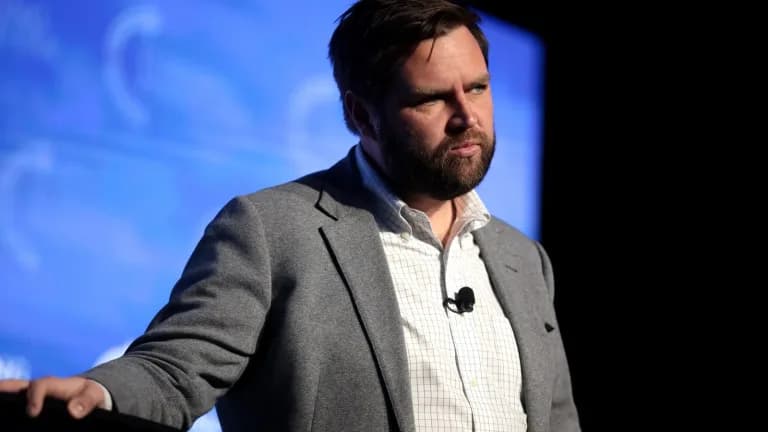 JD Vance in a gray blazer and checked shirt standing at a podium in front of a blue backdrop