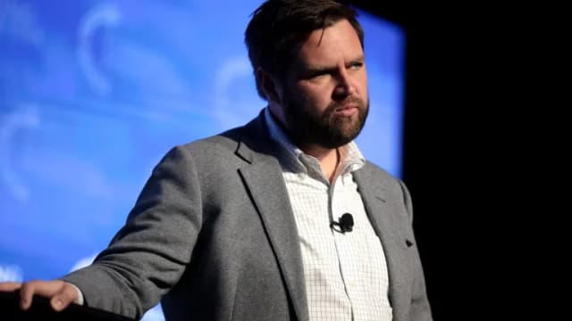 JD Vance in a gray blazer and checked shirt standing at a podium in front of a blue backdrop
