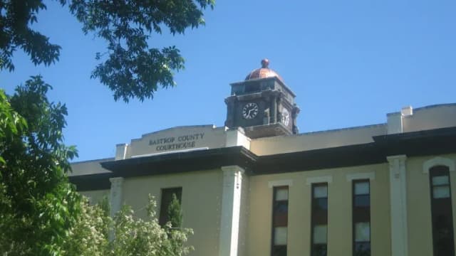 The upper floor and clock tower of the Bastrop County Courthouse framed by tree branches against a blue sky