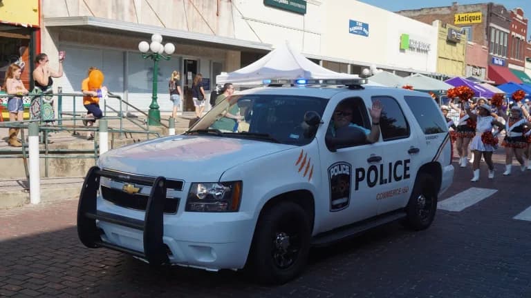 A white Commerce ISD Police Chevrolet Tahoe drives down a brick street during a small-town parade