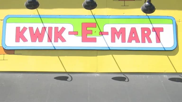 A red, green and white Kwik-E-Mart sign mounted on a bright yellow storefront wall above a gray awning