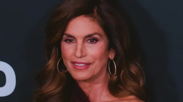 Cindy Crawford with long brown hair and gold hoop earrings at a red carpet event against a dark backdrop