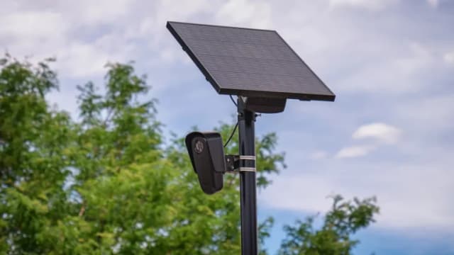 A solar-powered Flock Safety license plate reader camera mounted on a black pole in front of trees and sky