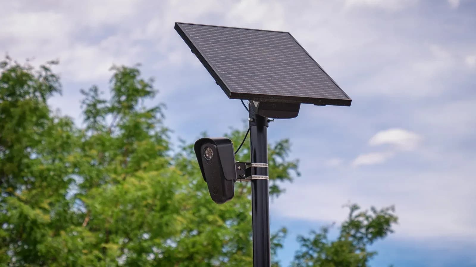 A solar-powered Flock Safety license plate reader camera mounted on a black pole in front of trees and sky