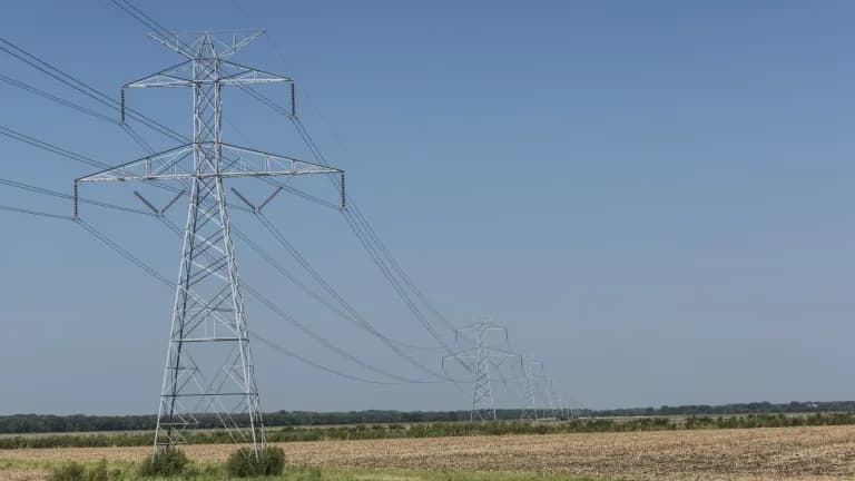 A row of steel electric transmission towers carrying power lines across flat farmland under a clear blue sky