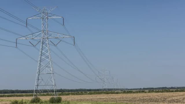 A row of steel electric transmission towers carrying power lines across flat farmland under a clear blue sky
