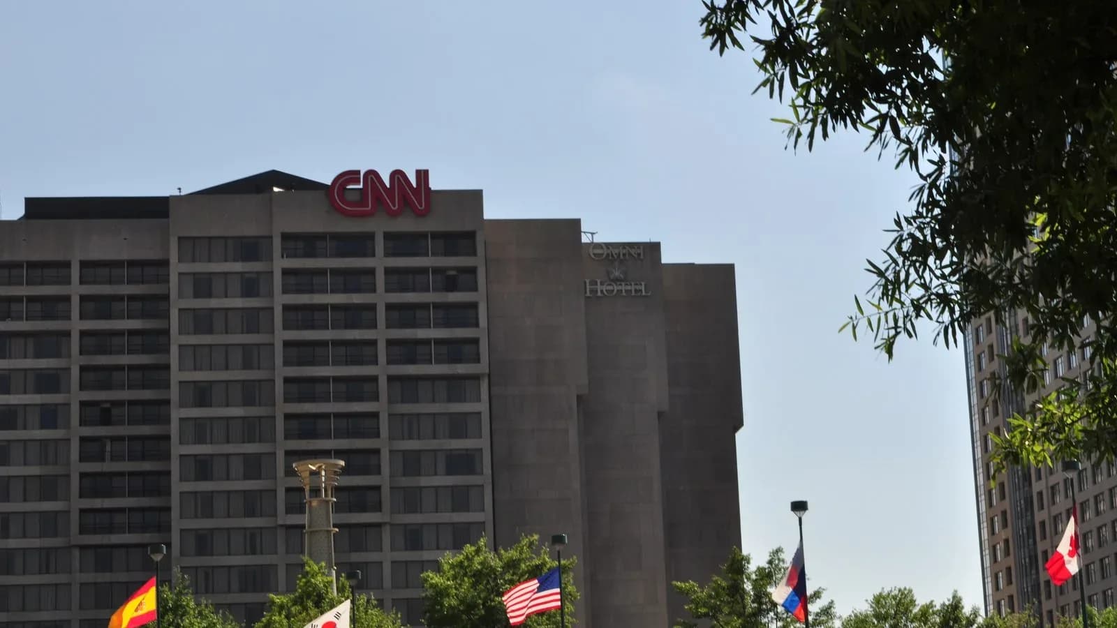 The red CNN letters atop the CNN Center building in downtown Atlanta under a clear sky, with flags and trees in the foreground