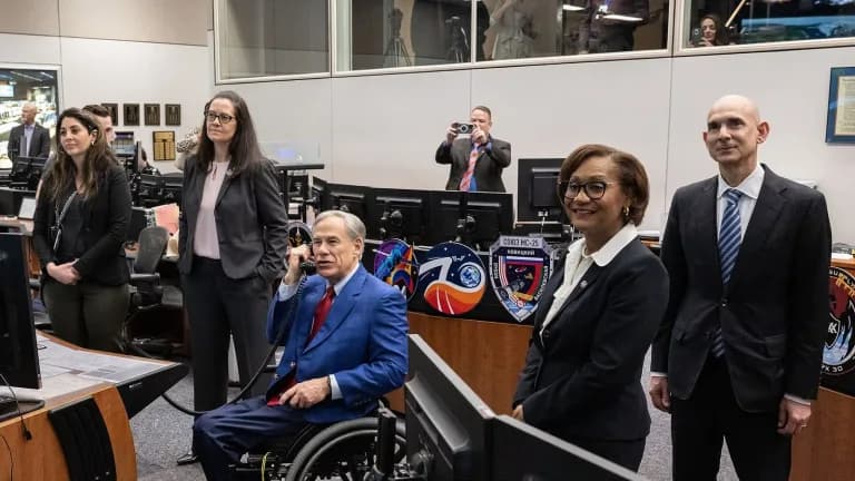 Gov. Greg Abbott, seated in a wheelchair and holding a phone to his ear, inside NASA’s Mission Control Center with officials standing beside him