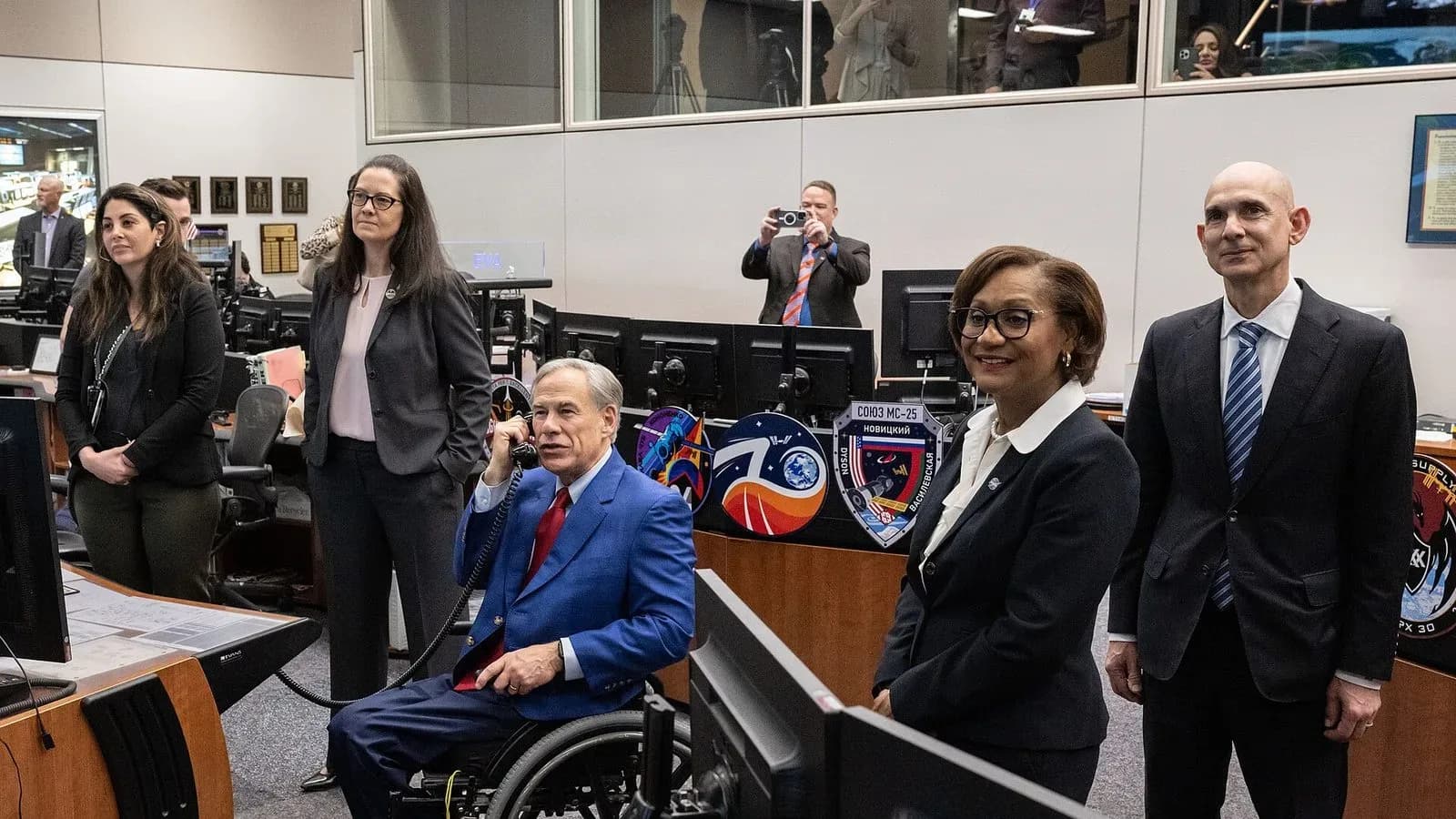 Gov. Greg Abbott, seated in a wheelchair and holding a phone to his ear, inside NASA’s Mission Control Center with officials standing beside him