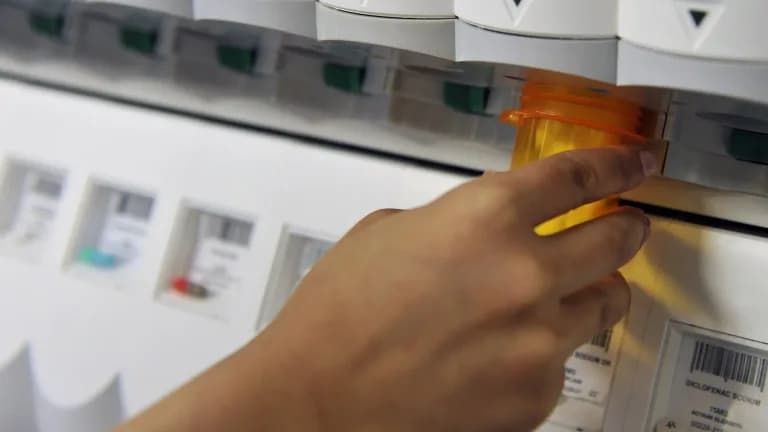 A hand pulling an orange prescription bottle from an automated medication dispenser in a pharmacy