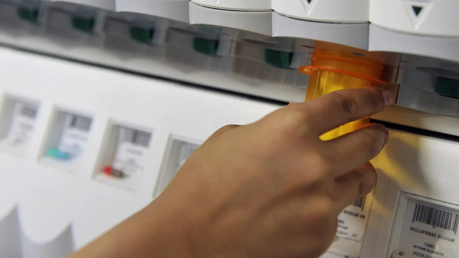 A hand pulling an orange prescription bottle from an automated medication dispenser in a pharmacy