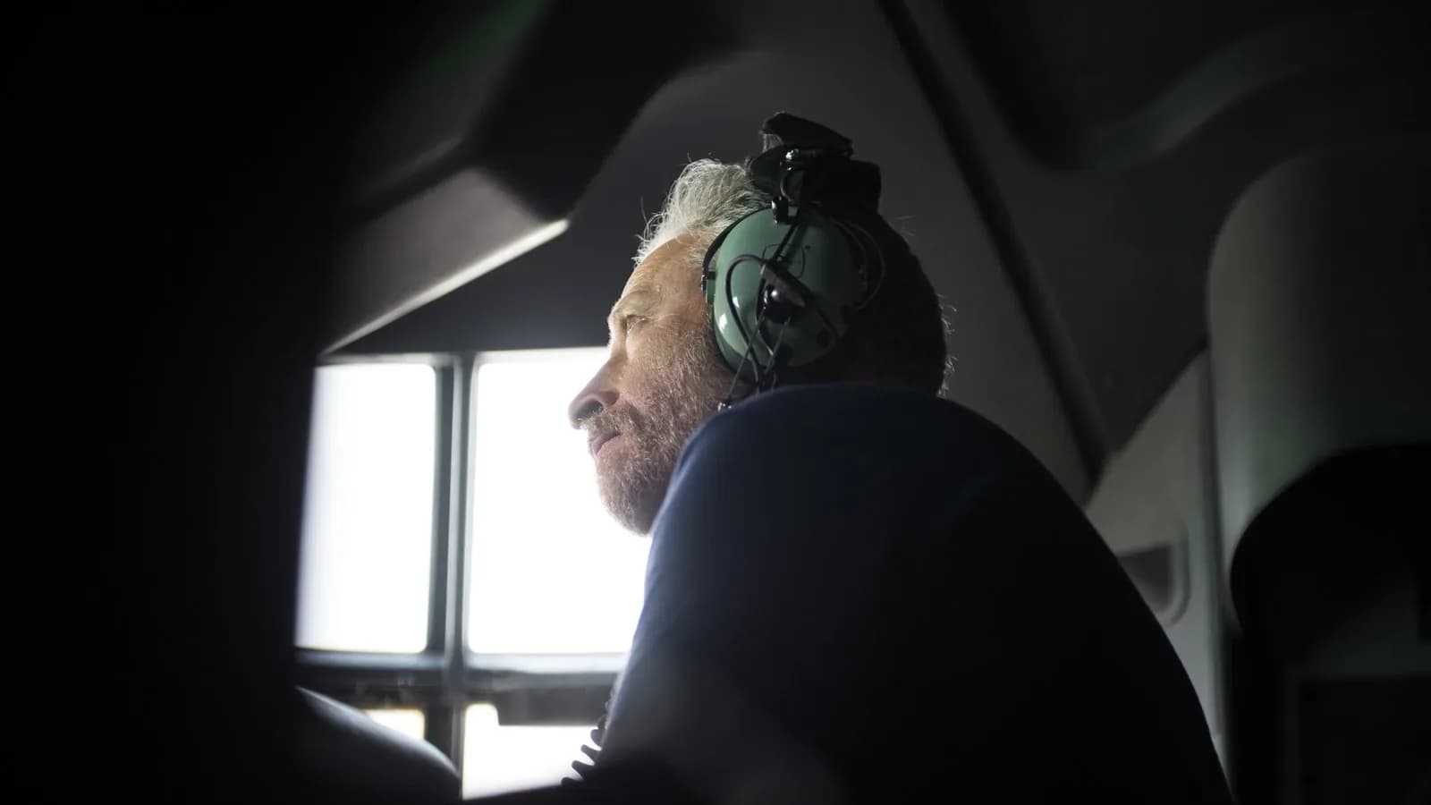 Jon Stewart, wearing a headset, looks out the cockpit window of a military aircraft in dim light