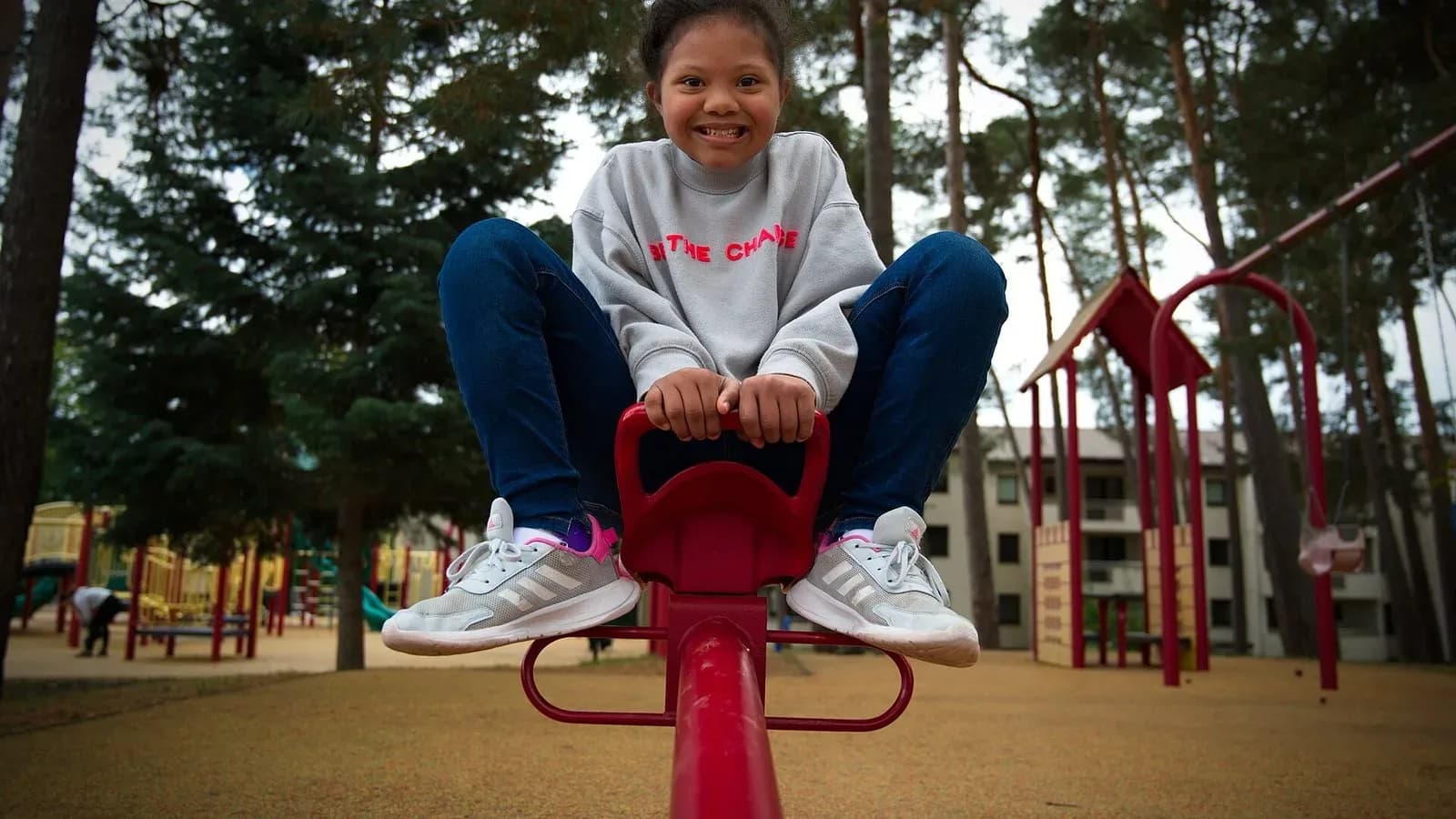A smiling young girl in a gray sweatshirt sits on a red teeter-totter at a playground