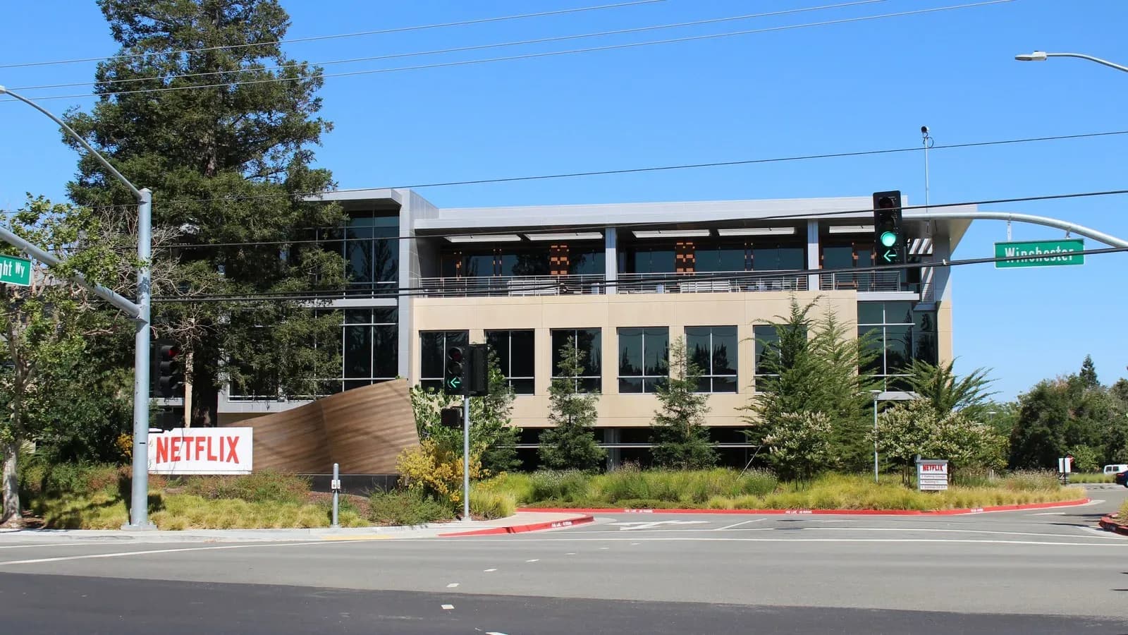A modern three-story office building behind a red-and-white Netflix sign at a landscaped street corner with traffic signals under a blue sky
