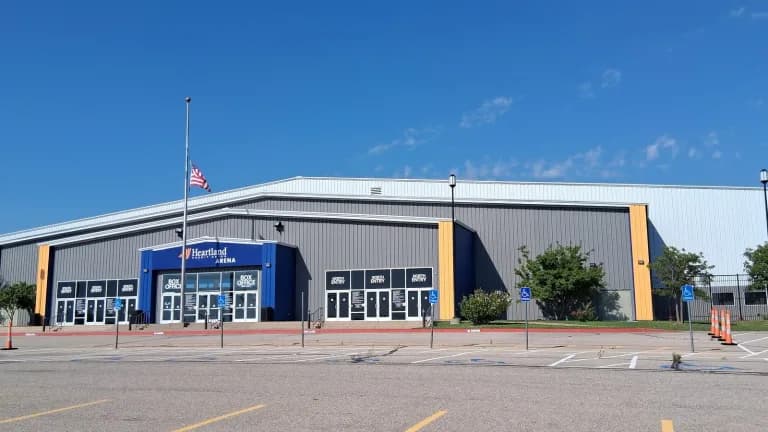 Exterior of a large gray arena with a blue Heartland Credit Union entrance, box office doors and an American flag on a pole under a clear blue sky