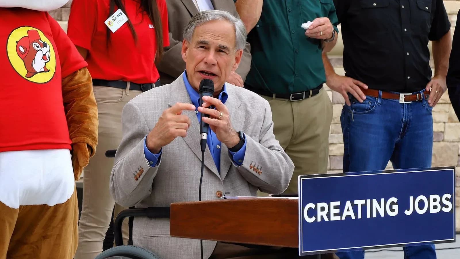 Gov. Greg Abbott, seated, speaks into a microphone at a podium with a sign reading Creating Jobs