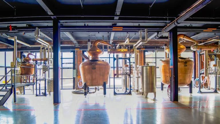 Copper pot stills of different sizes connected by pipes on a polished floor inside a bright distillery