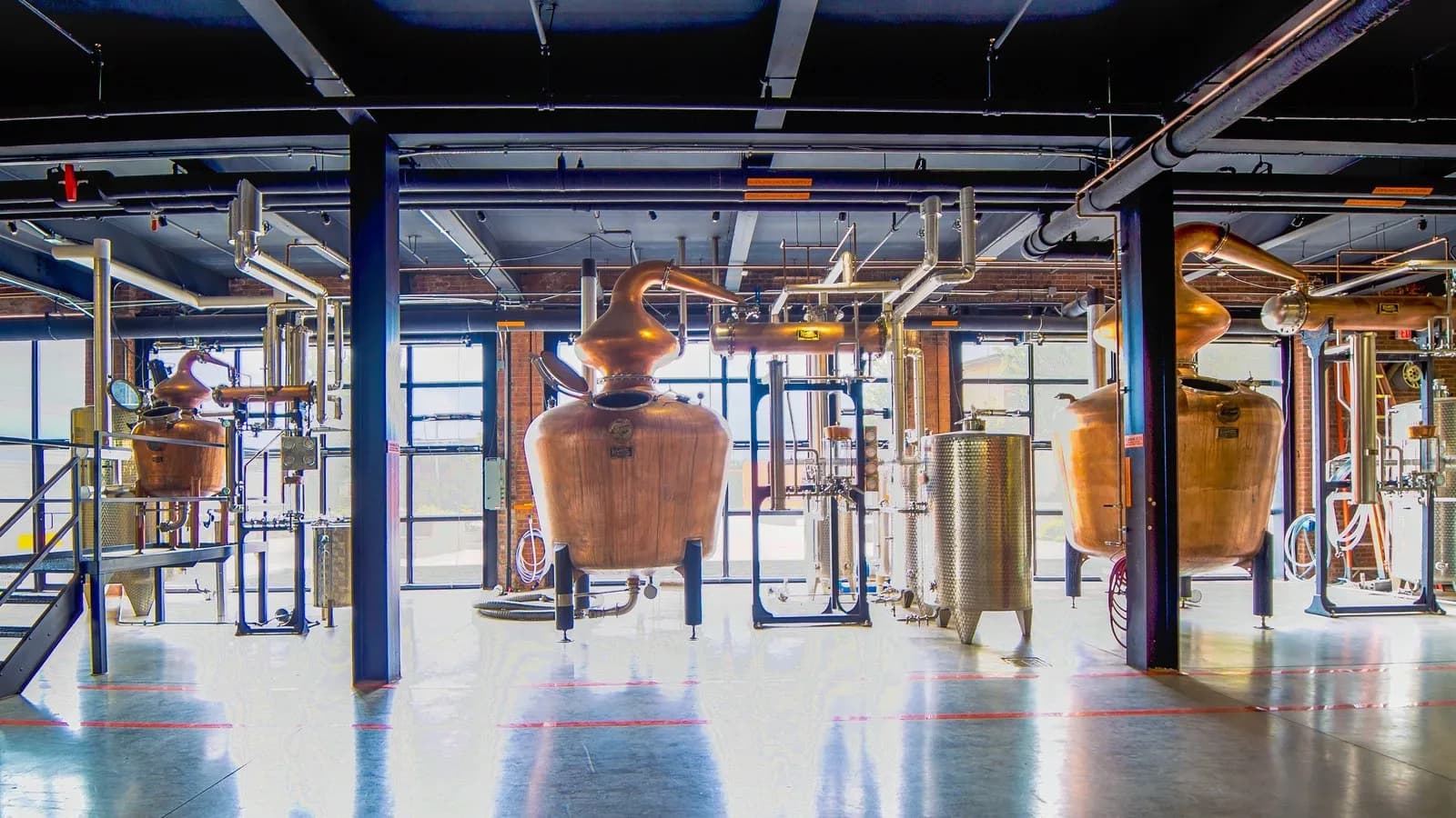 Copper pot stills of different sizes connected by pipes on a polished floor inside a bright distillery