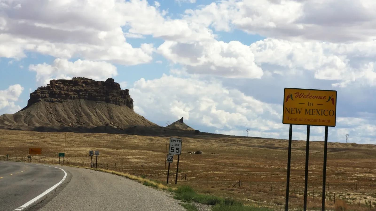 A yellow Welcome to New Mexico sign beside a two-lane highway with a flat-topped mesa and a cloudy sky behind it
