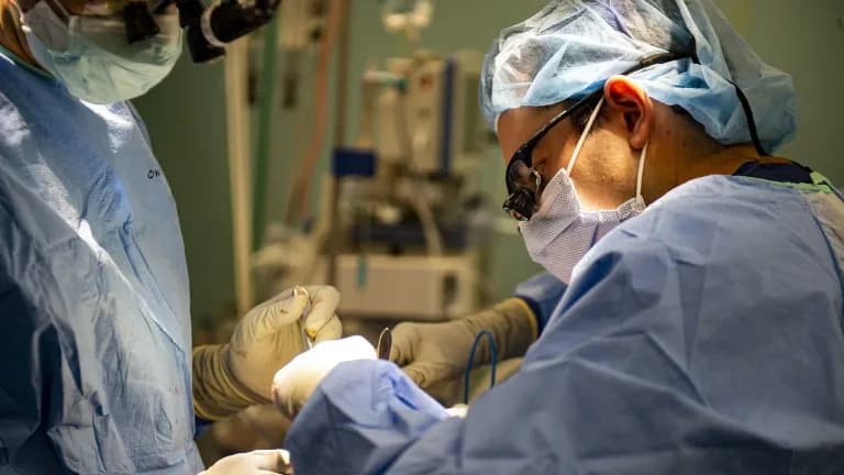 Two surgeons in masks, gowns and caps operate under bright lights in an operating room