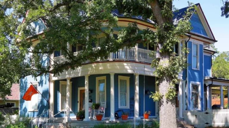 A blue two-story Victorian house with a wraparound porch beneath a large oak tree in Round Rock
