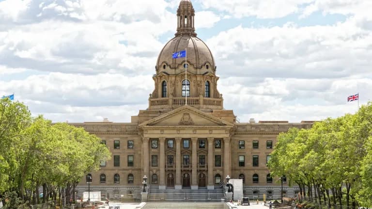 The domed sandstone Alberta Legislature Building in Edmonton behind trees and a plaza under a partly cloudy sky