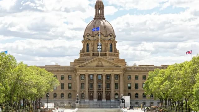 The domed sandstone Alberta Legislature Building in Edmonton behind trees and a plaza under a partly cloudy sky