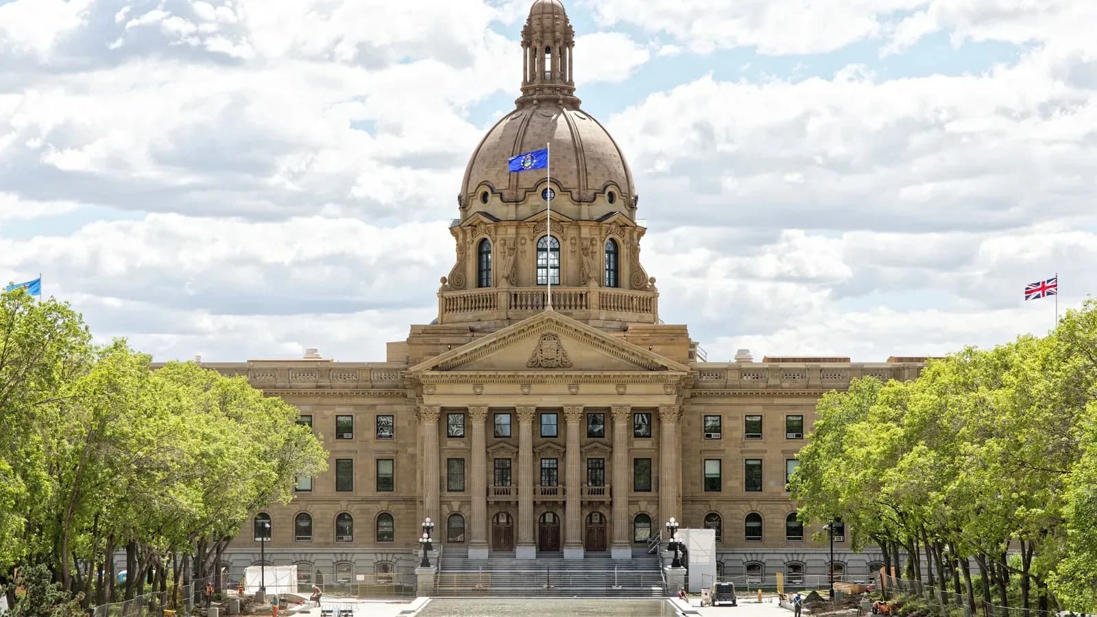 The domed sandstone Alberta Legislature Building in Edmonton behind trees and a plaza under a partly cloudy sky