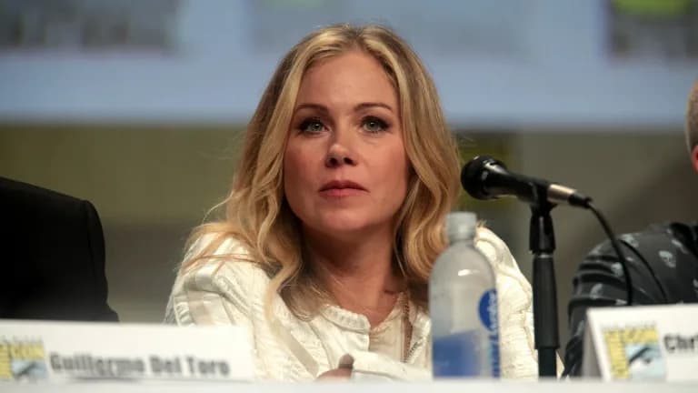 Christina Applegate, with long blond hair and a white top, seated behind a microphone at a convention panel