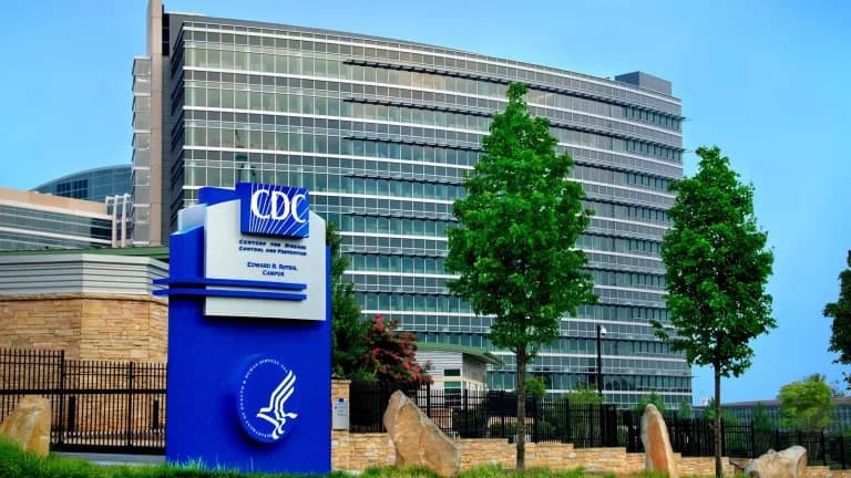 A blue CDC sign reading Centers for Disease Control and Prevention, Edward R. Roybal Campus, in front of a curved glass office building and trees