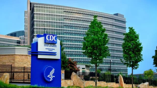 A blue CDC sign reading Centers for Disease Control and Prevention, Edward R. Roybal Campus, in front of a curved glass office building and trees