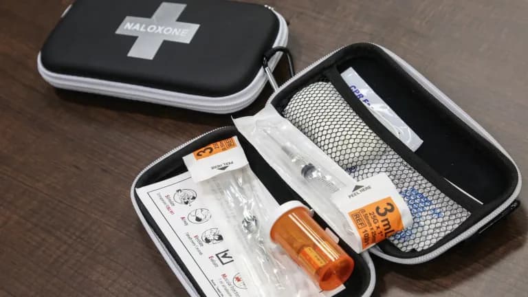 An open black naloxone kit on a wooden table showing a syringe, vials, gloves and instruction card