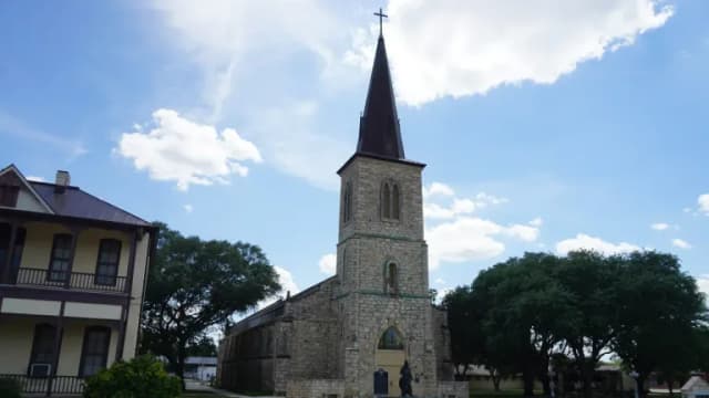 St. Louis Catholic Church in Castroville, Texas, a limestone Gothic Revival church with a tall dark spire and a statue of St. Louis in front, under a blue sky