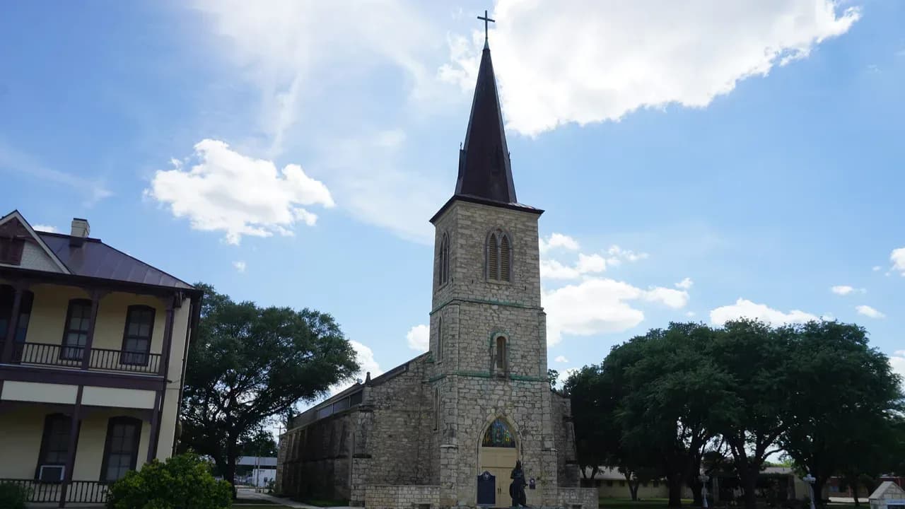 St. Louis Catholic Church in Castroville, Texas, a limestone Gothic Revival church with a tall dark spire and a statue of St. Louis in front, under a blue sky