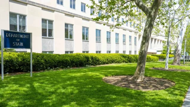 A blue Department of State sign on a green lawn in front of the pale stone Harry S. Truman Building, with trees and hedges
