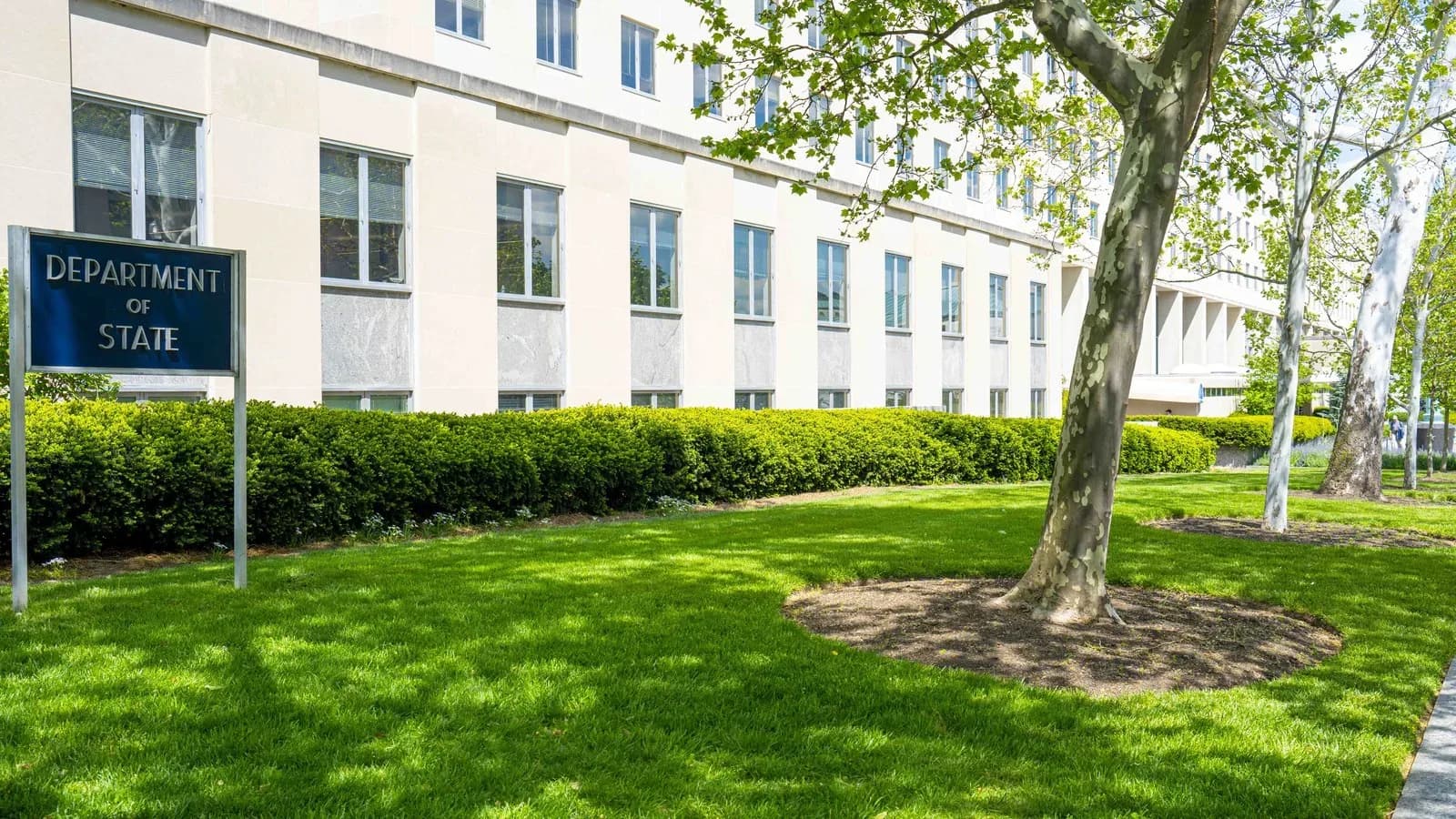 A blue Department of State sign on a green lawn in front of the pale stone Harry S. Truman Building, with trees and hedges
